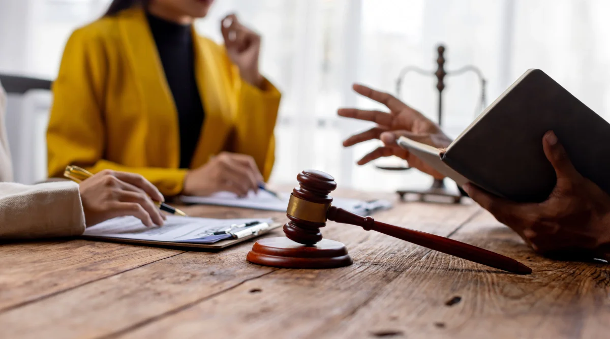 Close up of a judge's gavel resting on its stand while people talk in the background.